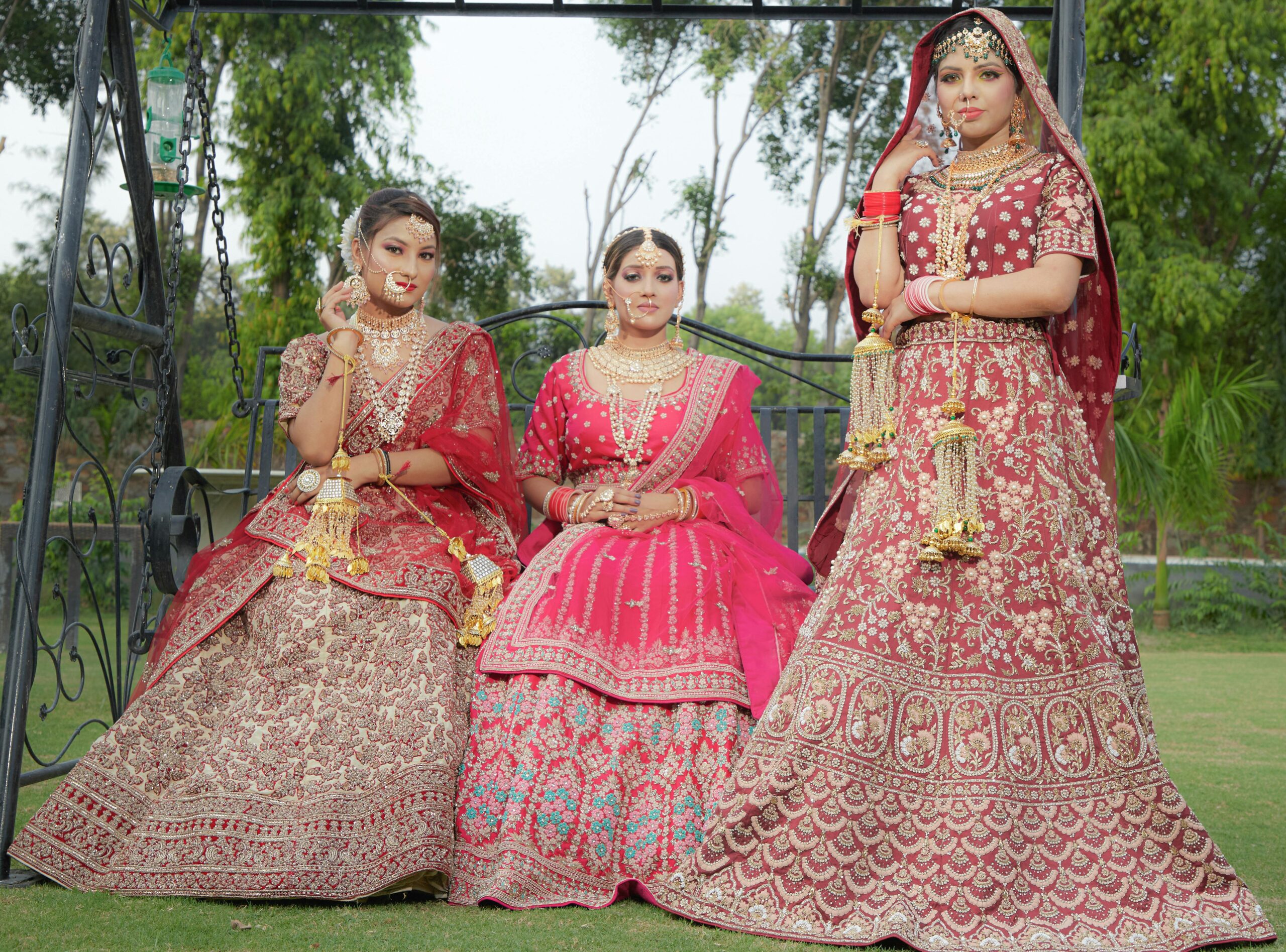 Three Indian women in exquisite traditional bridal lehengas sitting outdoors on a swing.