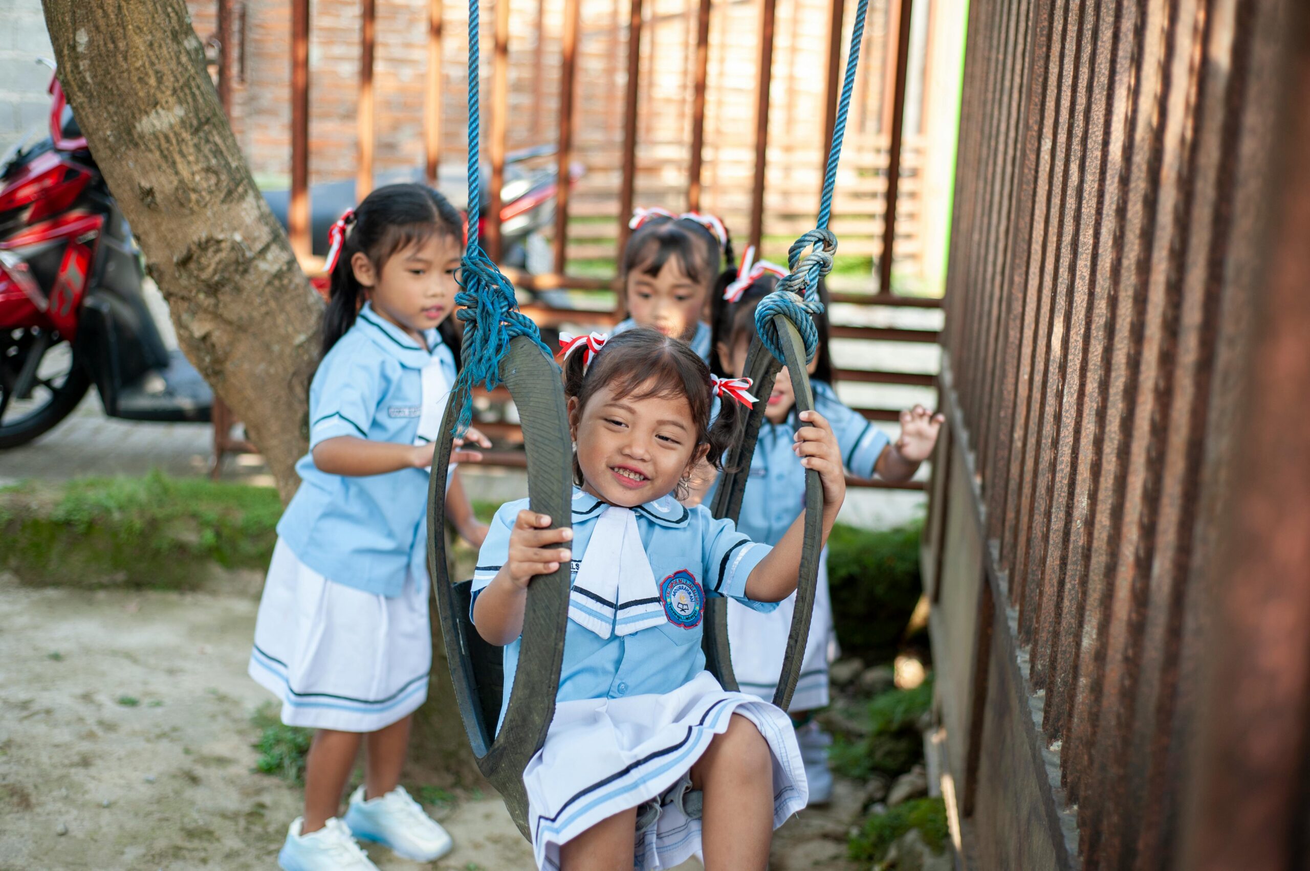 Three young girls play happily on swings in school uniforms, enjoying a sunny day outdoors.