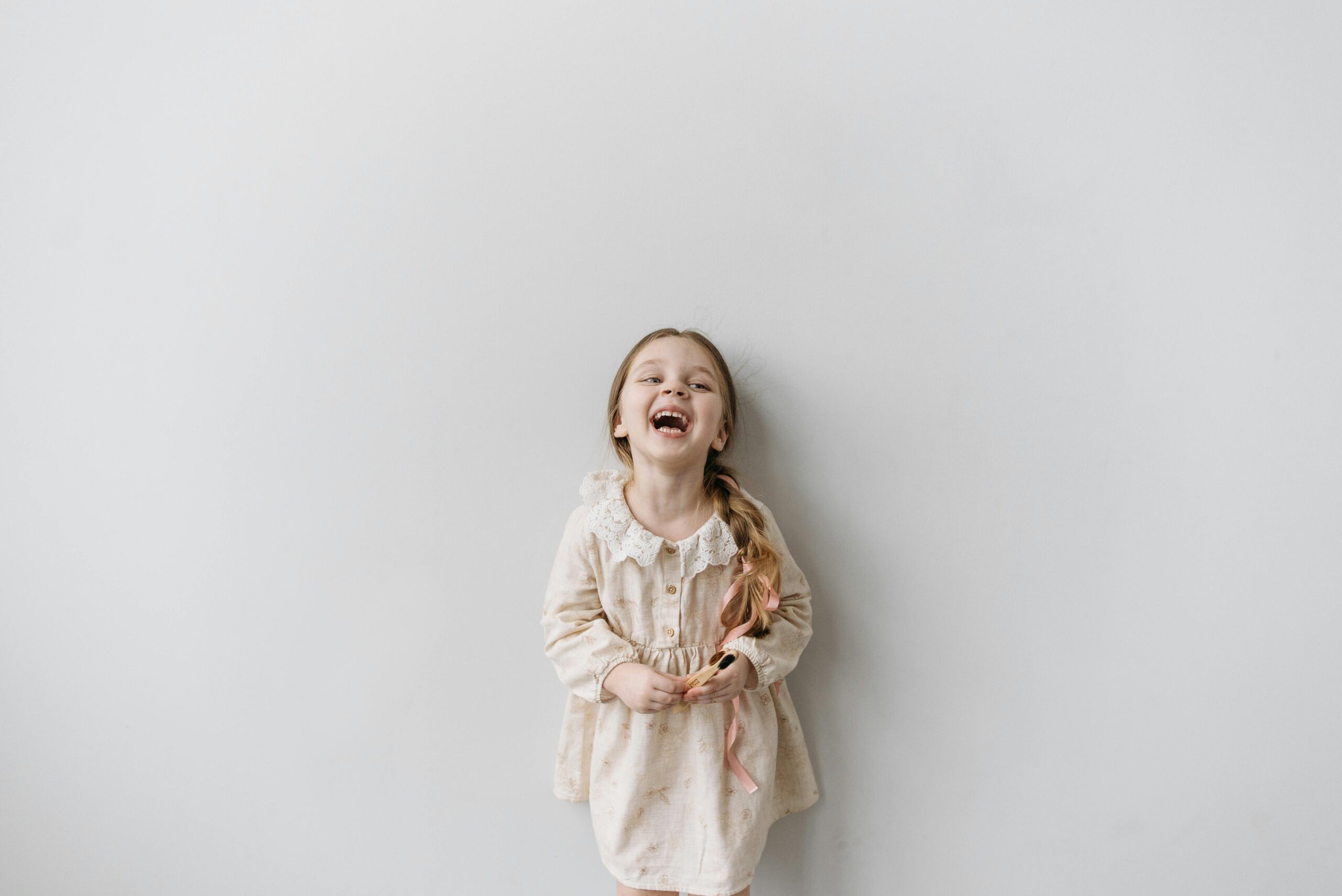 A happy young girl in a light dress laughing against a white wall.